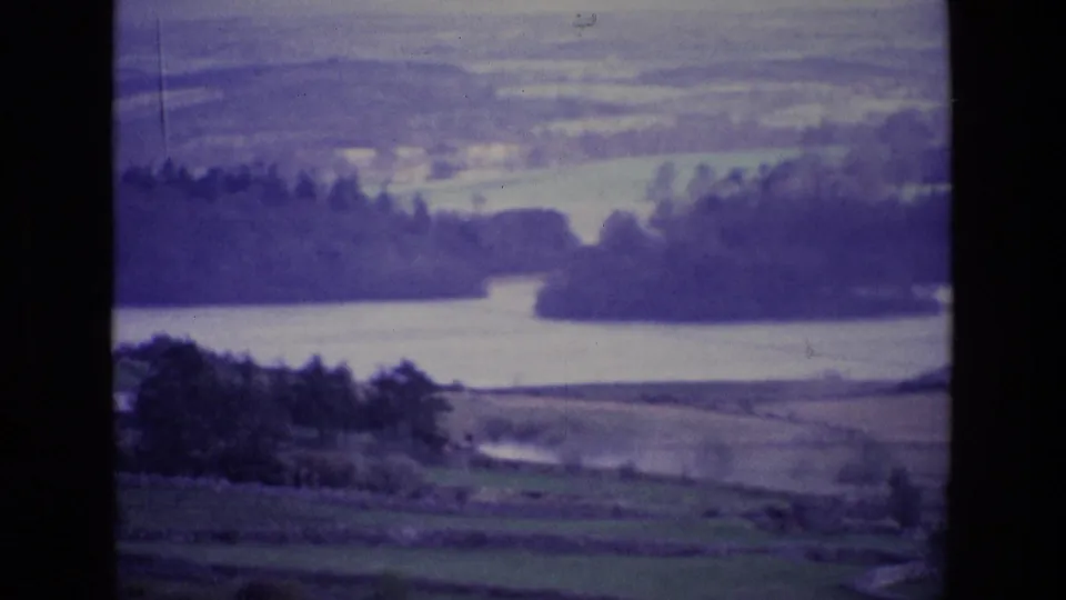 Aged 8mm Ireland frame of a lakeside rural landscape, with pasture fields, trees, water, and distant hills.