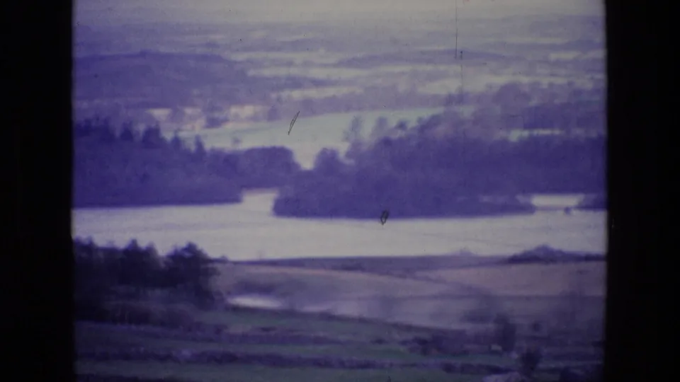 1969 8mm view of an Irish rural river valley with patchwork fields, dark tree lines, and soft distant hills.