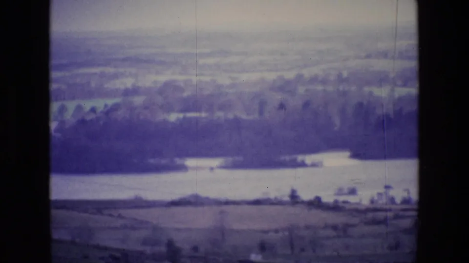 Late-1960s 8mm aerial frame over Ireland, showing countryside, wooded water edges, and a hazy distant landscape.