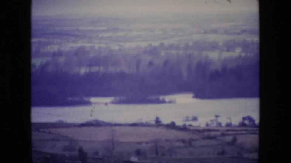 1969 Ireland 8mm frame showing a muted countryside panorama with lake water, woods, fields, scratches, and soft focus.