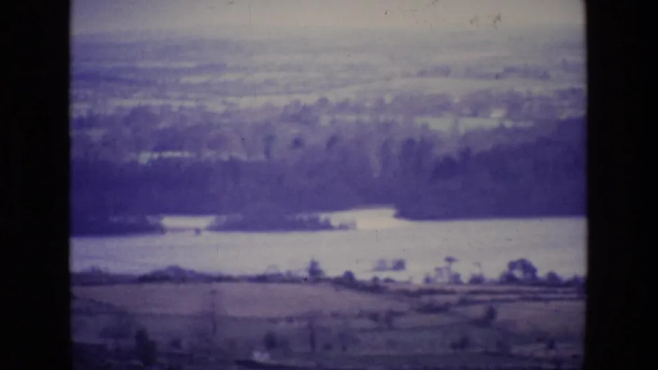 1969 Ireland home-movie frame showing a wide lakeside countryside view with fields, trees, haze, and dark film-frame borders.