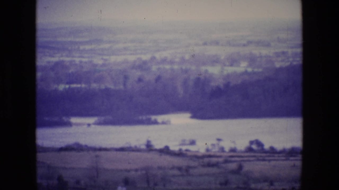 1969 Ireland 8mm frame showing a hazy lake, wooded shoreline, open fields, and distant rural landscape.