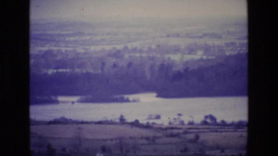 1969 Ireland 8mm frame showing a hazy lake, wooded shoreline, open fields, and distant rural landscape.