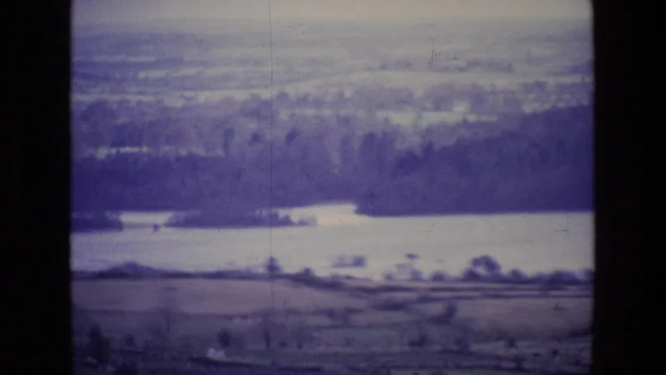 1969 Ireland 8mm frame with a lake, dense trees, fields, and hazy countryside under a pale sky.