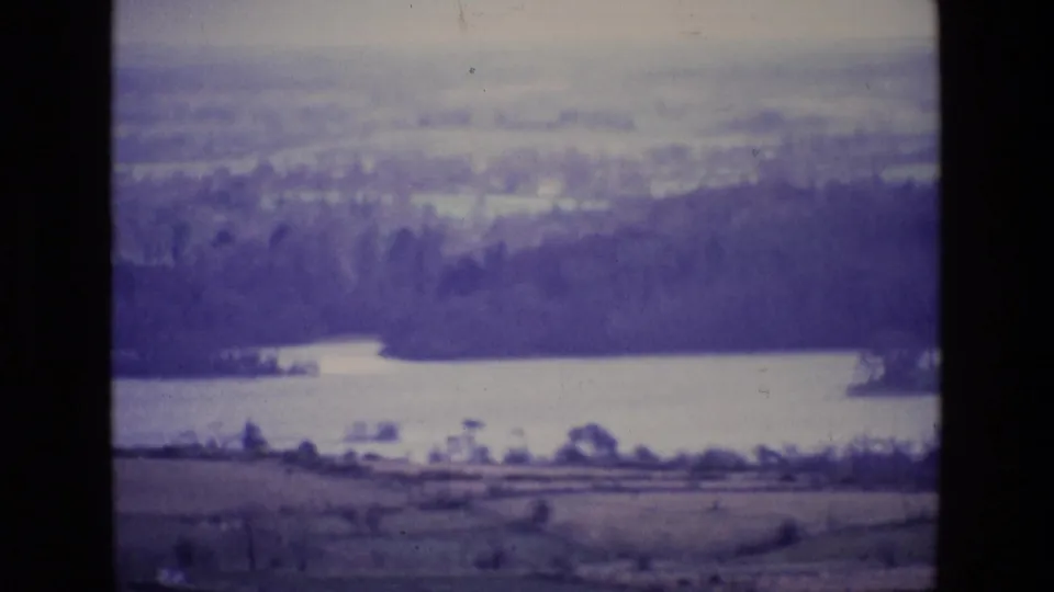 1969 Ireland 8mm frame of a lake and wooded shore with open land in the foreground and hazy distance beyond.