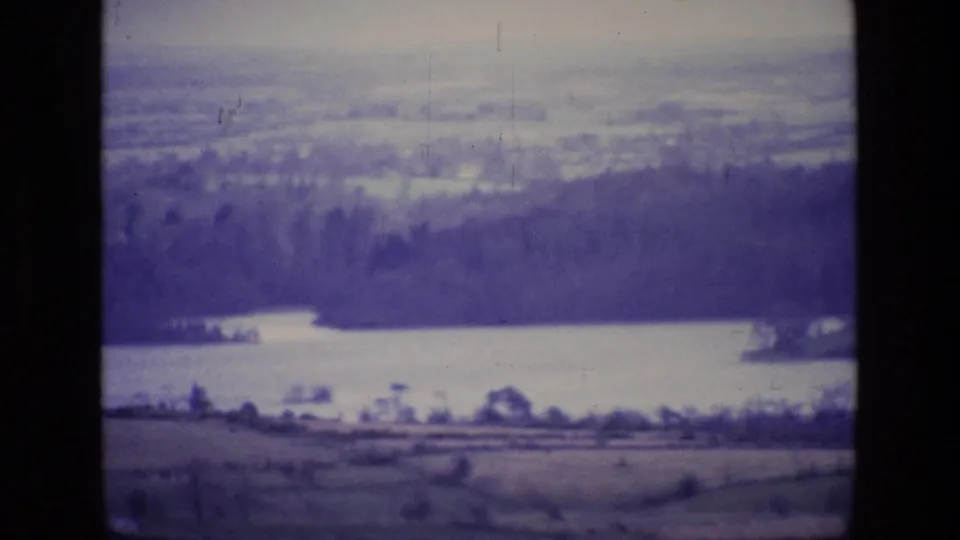 1969 8mm view in Ireland showing a lake, dark wooded ridge, fields, and distant rural horizon with a purple color cast.