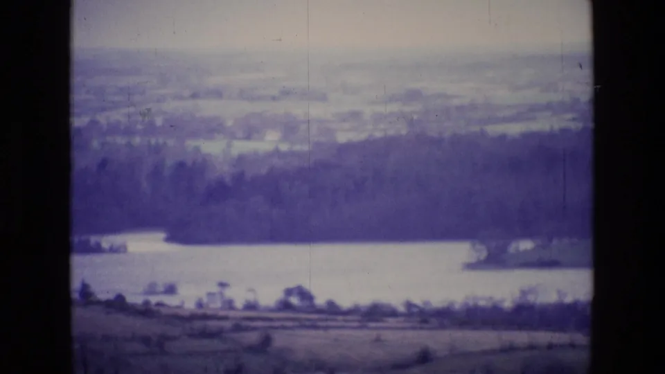 Soft 8mm Ireland landscape view from 1969, looking over fields toward a lake and wooded ridge under a hazy sky.