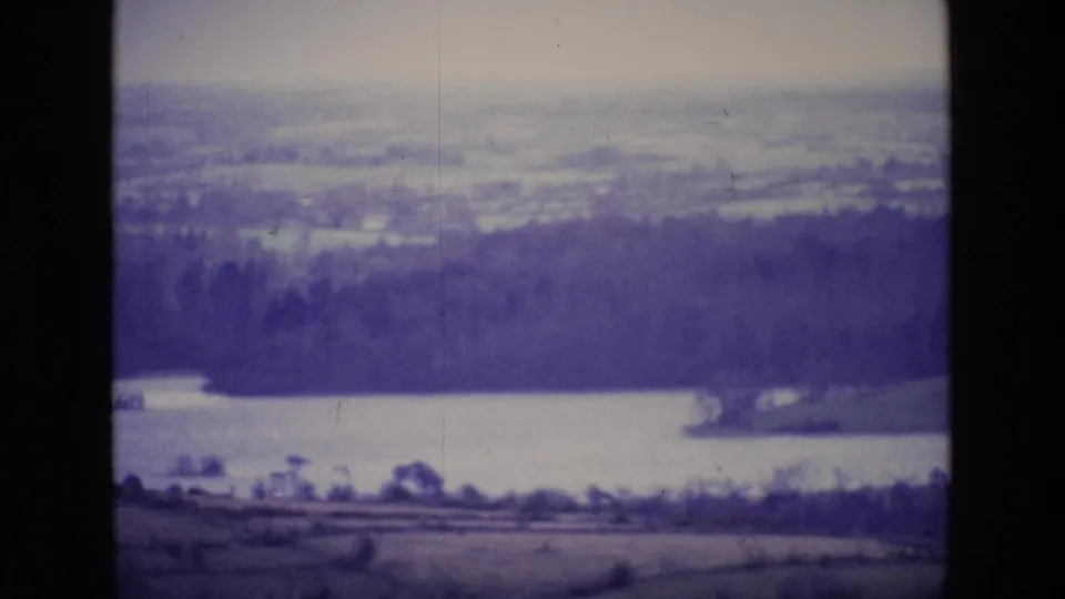Final 1969 8mm frame of an Irish lake landscape, with fields in front, wooded banks, and distant rural terrain.