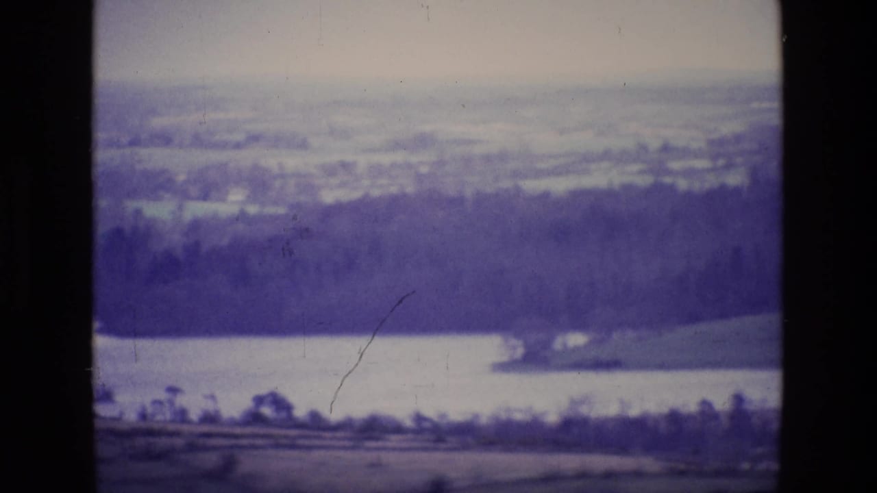 Distant lake below a dark wooded hillside with fields and a hazy sky.