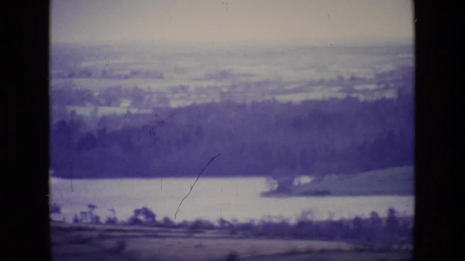 1969 Ireland 8mm frame showing a distant lake, dark forested hillside, fields, and a pale hazy horizon.