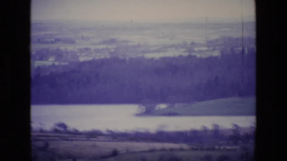 1969 Ireland 8mm frame showing a lake at the foot of a wooded hill with fields in the foreground.