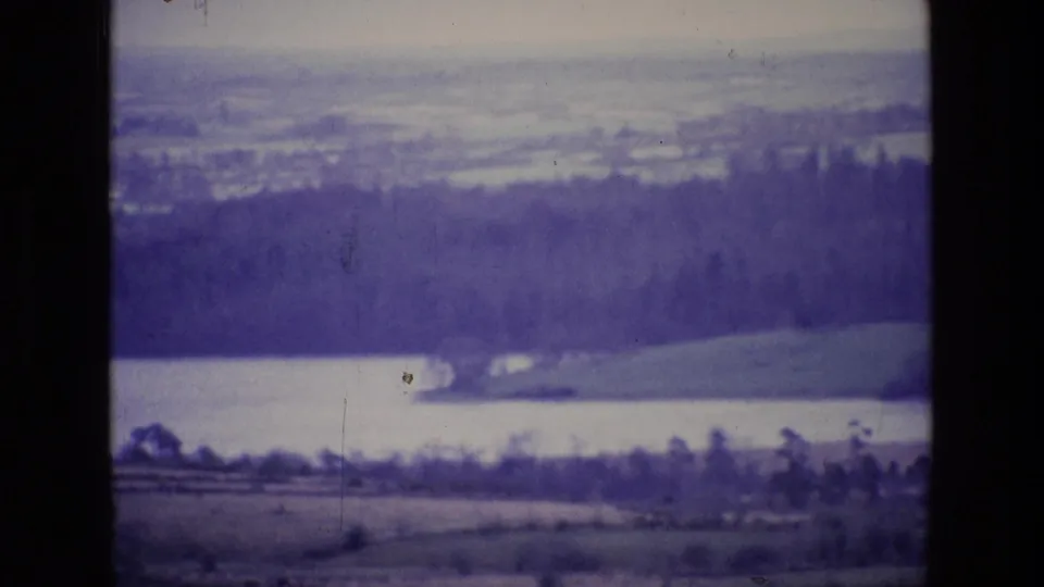1969 Ireland home-movie frame with a lake below a dark forested hillside and distant rural fields.