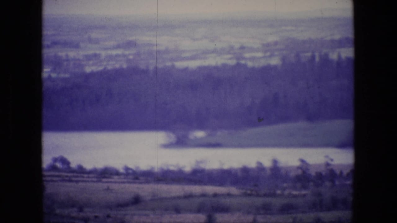 1969 Ireland 8mm view of a pale lake, wooded slopes, distant fields and dark film gate borders with a purple cast.