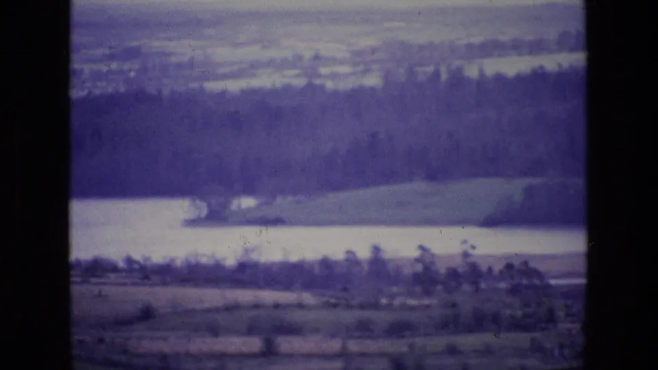 Soft 8mm frame of an Irish lake landscape, with wooded hills, pale water and rural fields under aged purple color shift.