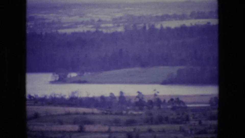 1969 Irish home-movie frame showing a lake below dense trees and rolling farmland, with soft focus and purple film cast.