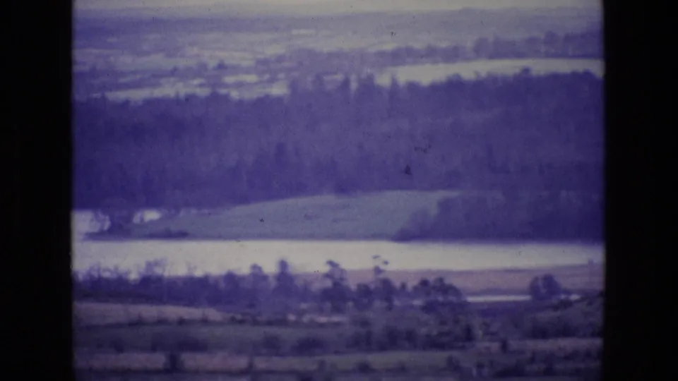 Vintage 8mm view from Ireland in 1969, showing pale lake water, tree-covered hills and open fields within dark film borders.
