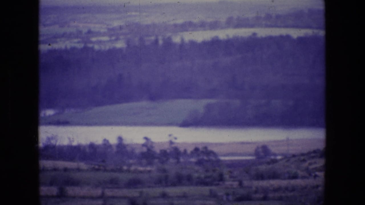 1969 8mm home-movie frame from Ireland showing layered fields, a pale lake or river, wooded bands, and distant hills with purple film cast.