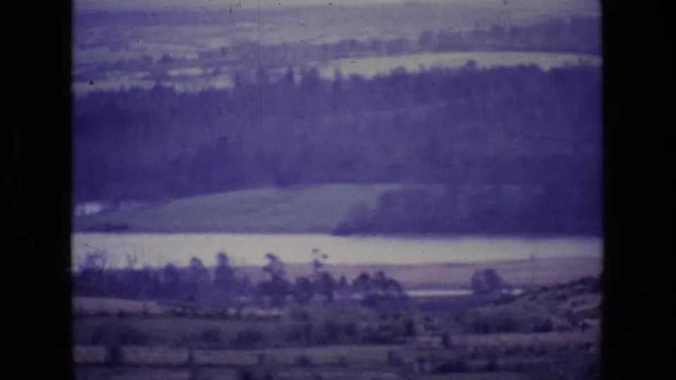 1969 8mm home-movie frame from Ireland showing layered fields, a pale lake or river, wooded bands, and distant hills with purple film cast.