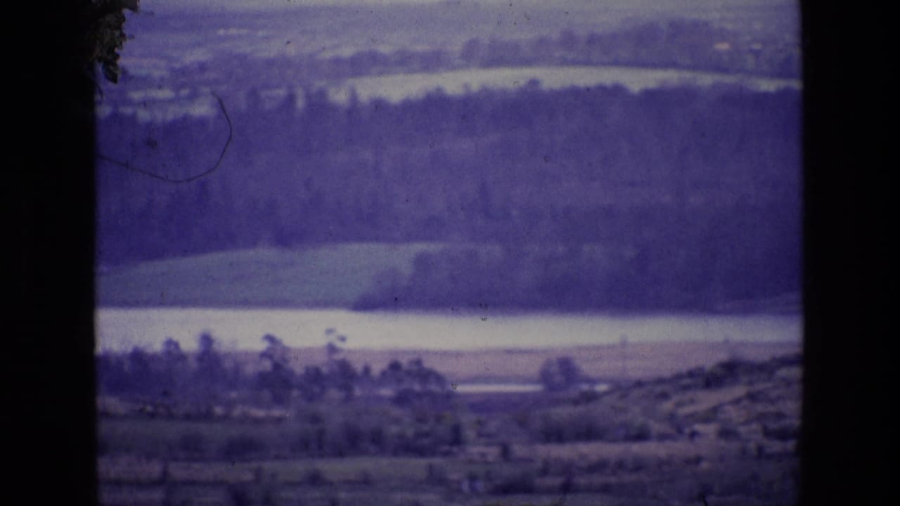 Wide rural landscape with pale water, wooded bands, hills, and a dark central obstruction.