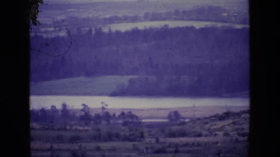 1969 Ireland landscape frame showing a lake or river below wooded hills, with a dark vertical foreground strip dividing the view.