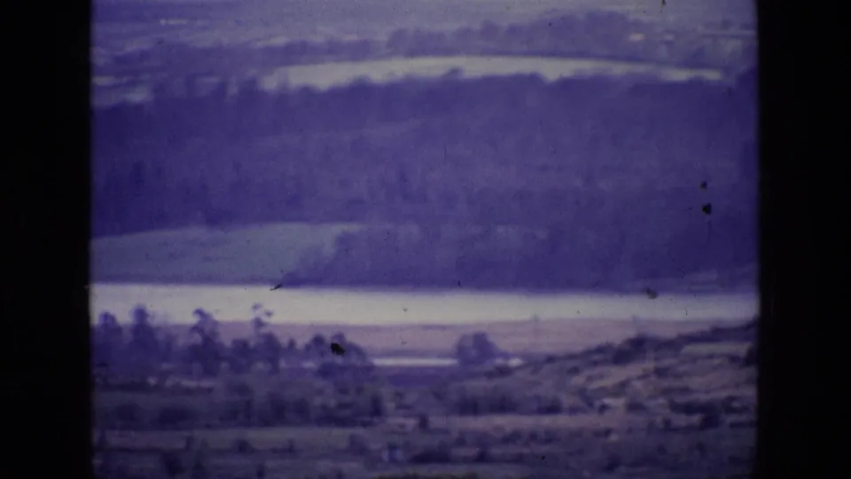 1969 8mm frame from Ireland with soft-focus fields, pale water, treeline, and distant hills under a purple-tinted film image.