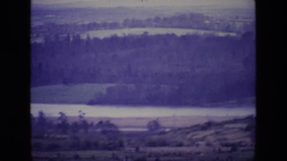 Late-1960s Ireland home-movie frame showing layered countryside, pale water, trees, and hills with visible 8mm grain and color shift.