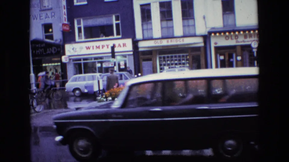 1969 Ireland town street frame with a dark car passing across the foreground, wet pavement, shopfronts, pedestrians, and parked traffic.