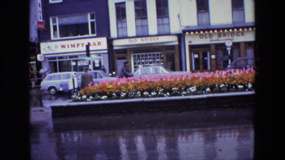 1969 Ireland home-movie frame showing a rainy town street, flower bed, storefront signs, pedestrians, and cars near an intersection.
