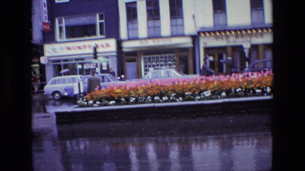 1969 Ireland street view with a colorful flower bed, wet pavement, parked cars, pedestrians, and Wimpy Bar and Old Bridge storefront signs.