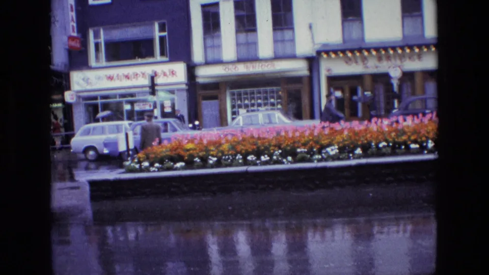 1969 Ireland street view with a colorful flower bed, wet pavement, parked cars, pedestrians, and Wimpy Bar and Old Bridge storefront signs.