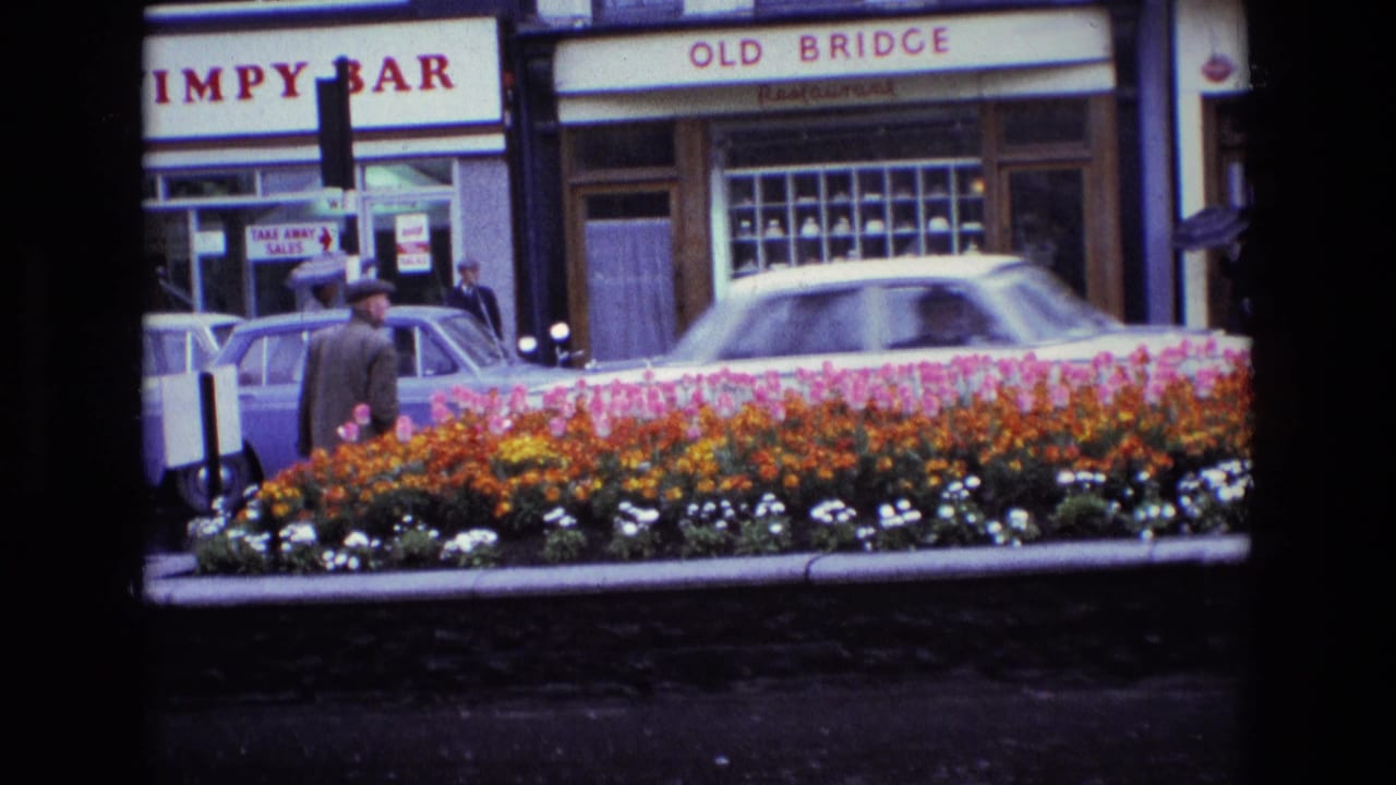 1969 Ireland street frame with a man in a dark coat near flowers, parked cars, and readable Wimpy Bar and Old Bridge Restaurant signage.