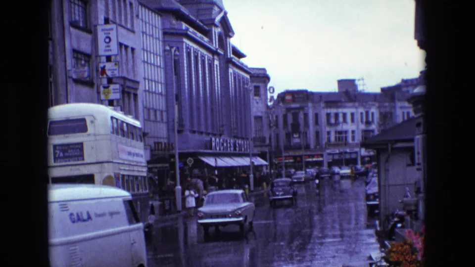 Rain-slick Irish downtown street from 1969 with vintage cars, a double-decker bus, pedestrians, and visible commercial signs.