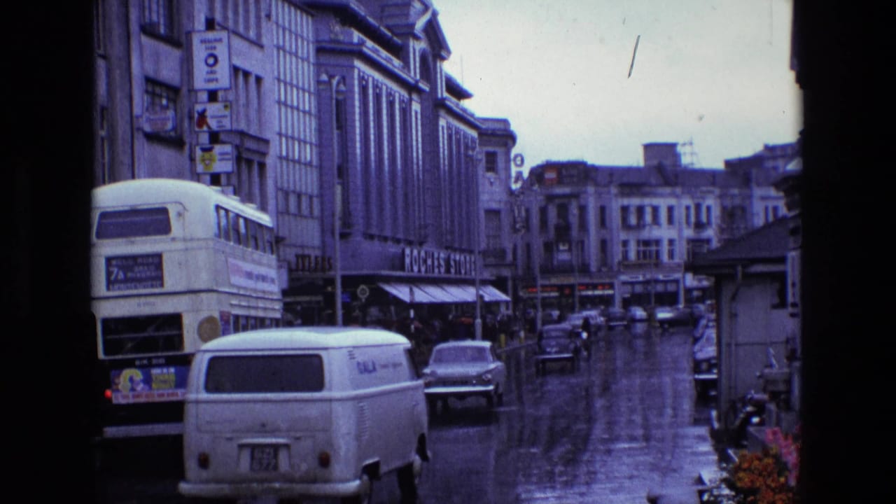 1969 Ireland street view showing rain-wet pavement, a double-decker bus, a white van, cars, and commercial buildings from a fixed home-movie angle.