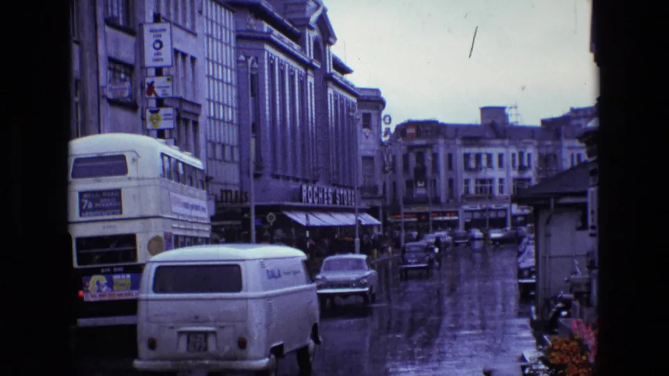 1969 Ireland street view showing rain-wet pavement, a double-decker bus, a white van, cars, and commercial buildings from a fixed home-movie angle.