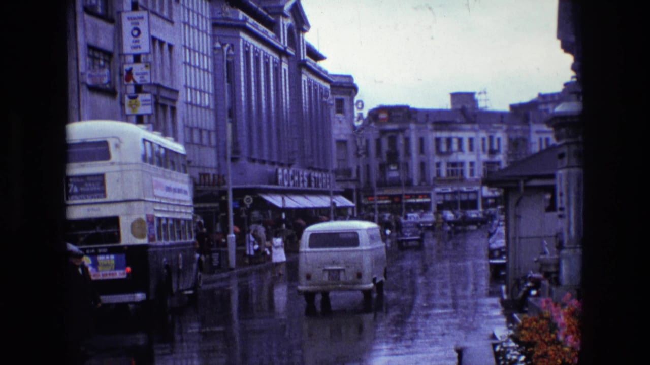 1969 Ireland rainy street scene with a white van in the roadway, a double-decker bus at left, pedestrians near storefronts, and reflective pavement.