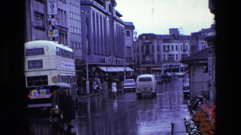 1969 Ireland city street showing umbrella-carrying pedestrians in the foreground, a double-decker bus, cars, storefronts, and rain-reflective pavement.