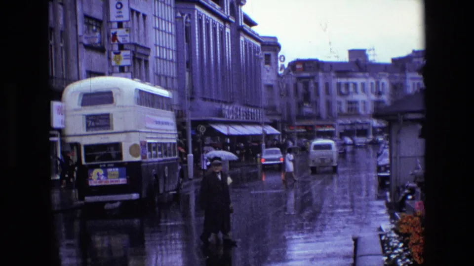 1969 Ireland rainy urban view with a pedestrian holding an umbrella in the foreground, a double-decker bus, moving cars, storefronts, and wet reflections.