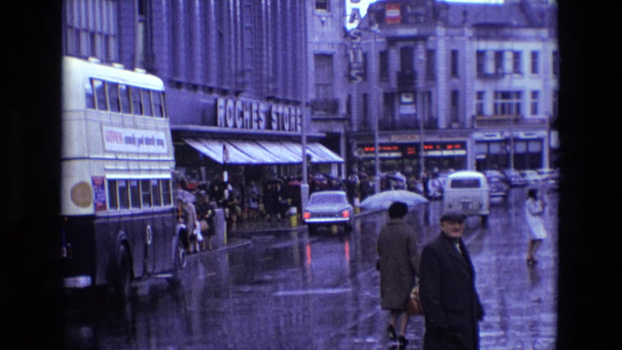 Rainy 1969 Ireland street with a bus, pedestrians, vintage cars, shopfronts and bright reflections on wet pavement.
