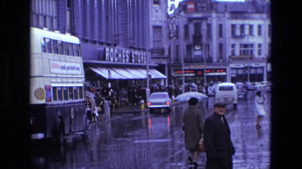 Rainy 1969 Ireland street with a bus, pedestrians, vintage cars, shopfronts and bright reflections on wet pavement.