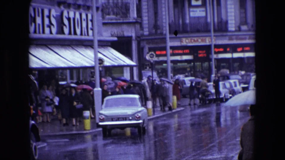 1969 Ireland rainy shopping street with umbrella-carrying pedestrians, older cars, storefront signage and reflective pavement.