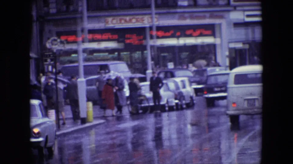Rainy 1969 Ireland street view showing pedestrians at the curb, older cars, a white van and shop signs across the road.