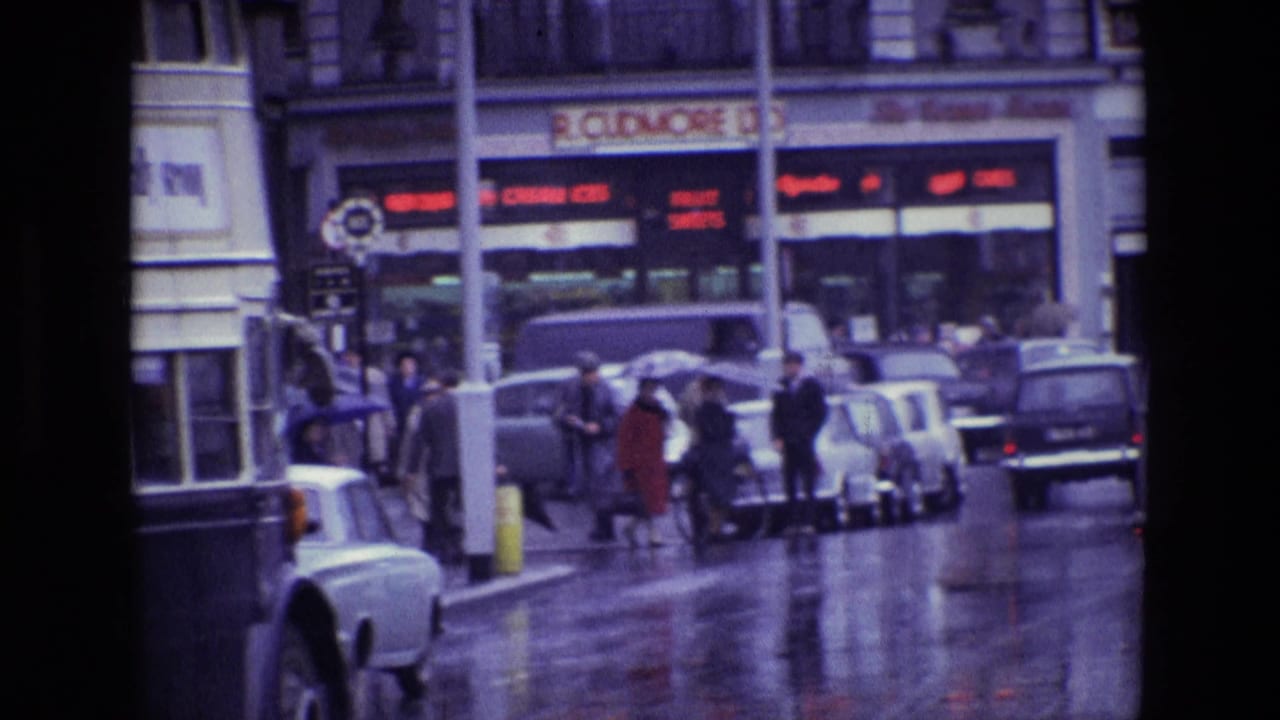 1969 Ireland street view with wet pavement, pedestrians, parked cars, and a double-decker bus passing a commercial block.