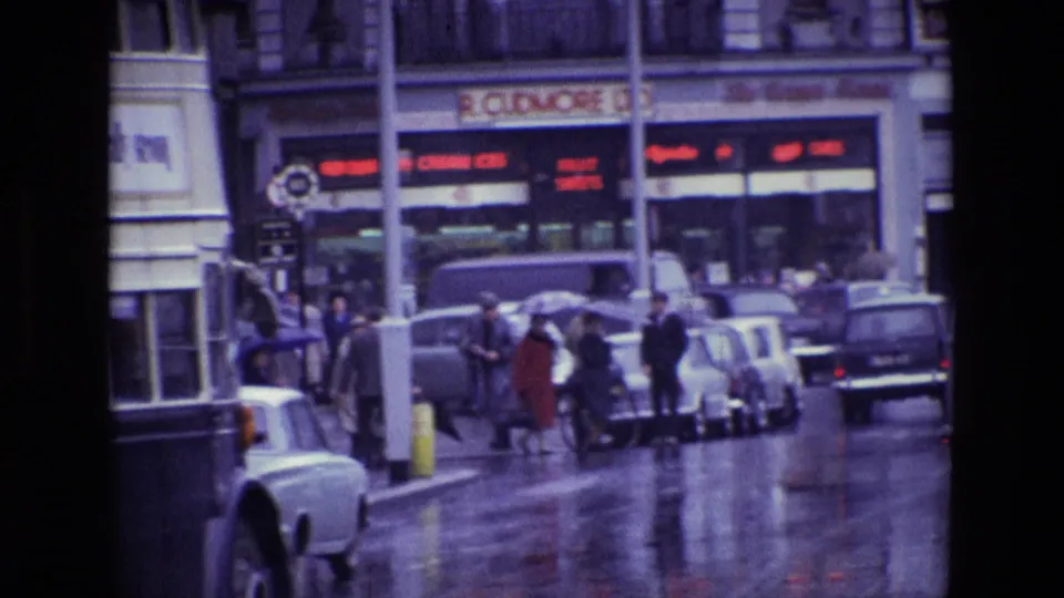 1969 Ireland street view with wet pavement, pedestrians, parked cars, and a double-decker bus passing a commercial block.