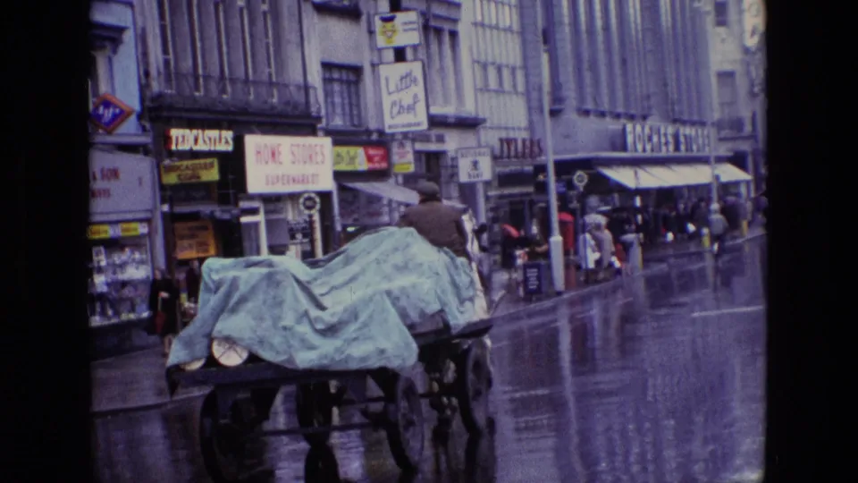 Rear view of a covered horse-drawn cart traveling through a rainy 1969 Irish shopping street with pedestrians nearby.