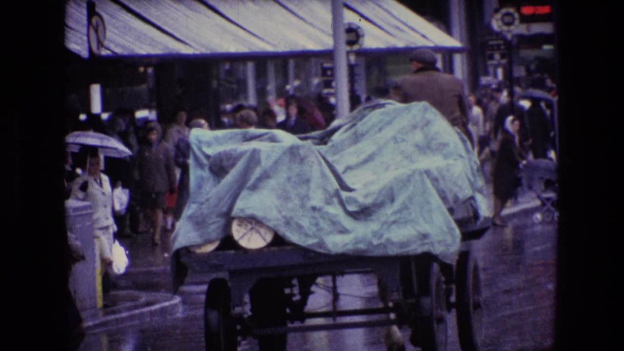 A covered cart or truck passes through a rainy Irish city street in 1969, with pedestrians, umbrellas, wet pavement, and shop awnings visible.