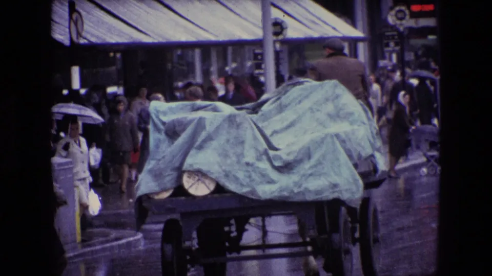 A covered cart or truck passes through a rainy Irish city street in 1969, with pedestrians, umbrellas, wet pavement, and shop awnings visible.