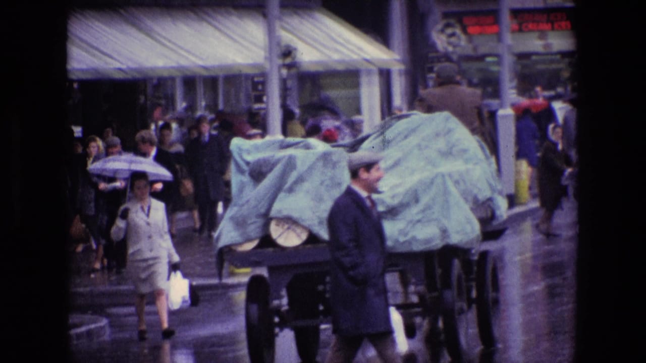 Pedestrians and a man in a dark coat pass a covered vehicle on a wet street.