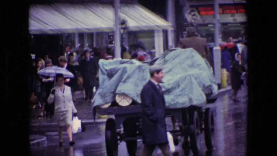 In a 1969 Irish city street scene, pedestrians with umbrellas move around a covered vehicle while a man in a dark coat crosses the foreground.