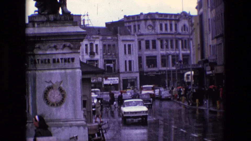 The Father Mathew monument appears beside a rainy Irish city street, with pedestrians, older cars, and commercial buildings in the background.
