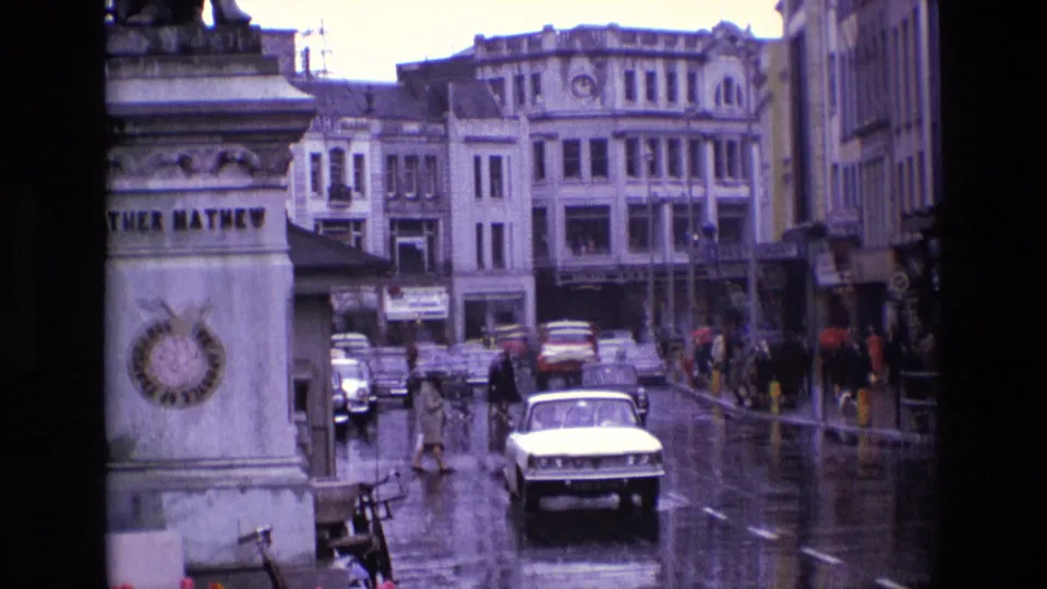 A white 1960s car approaches on a wet Irish city street near the Father Mathew monument, with pedestrians and storefronts beyond.